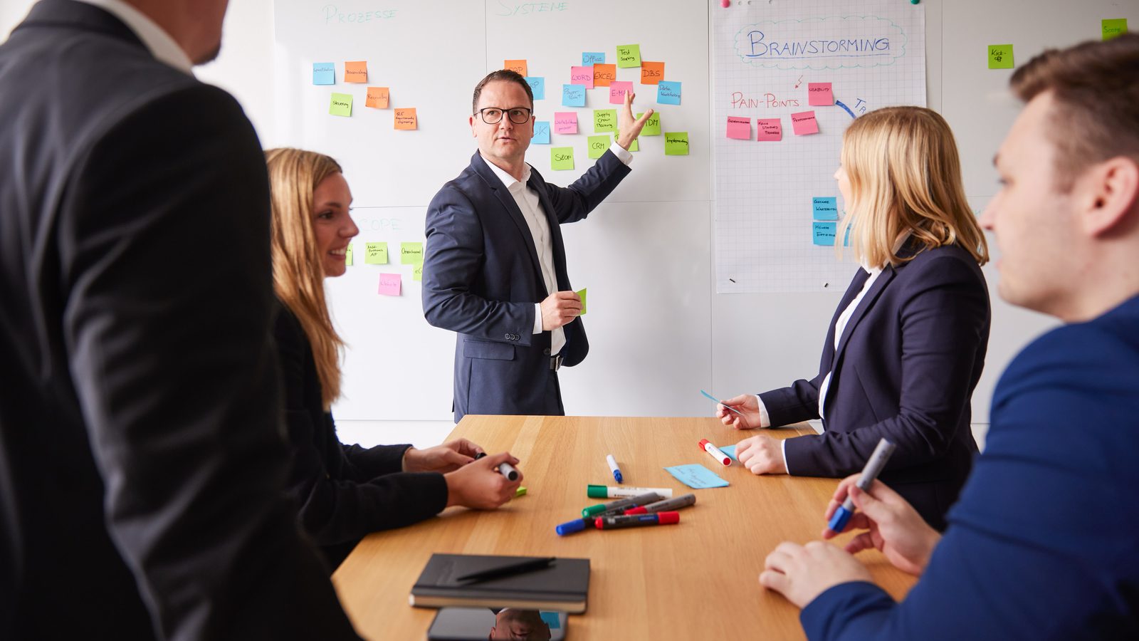 Workshop bei ReqPOOL: Ein Berater erläutert an einer Wand mit Haftnotizen die nächsten Entscheidungen, das Team sitzt am Tisch.