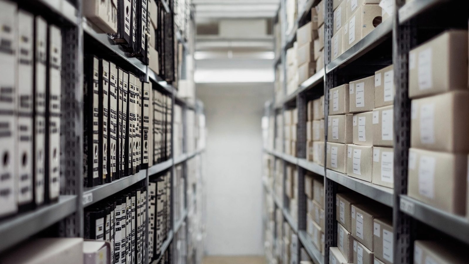 Blick in einen Archivgang: links Regale mit Aktenordnern, rechts Regale mit beschrifteten Archivkartons, am Ende des Gangs helles Licht.