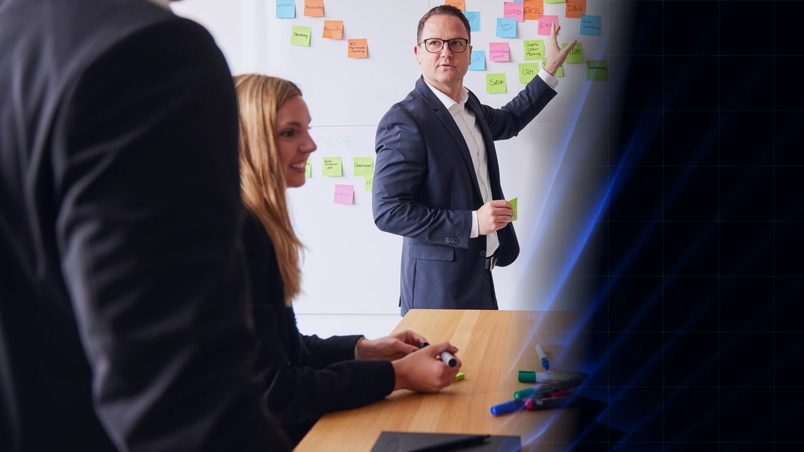Workshop bei ReqPOOL: Ein Berater zeigt auf eine Wand mit bunten Haftnotizen, eine Kollegin macht sich am Tisch Notizen; rechts geht das Bild in das blaue Formenmuster der ReqPOOL-Website über.