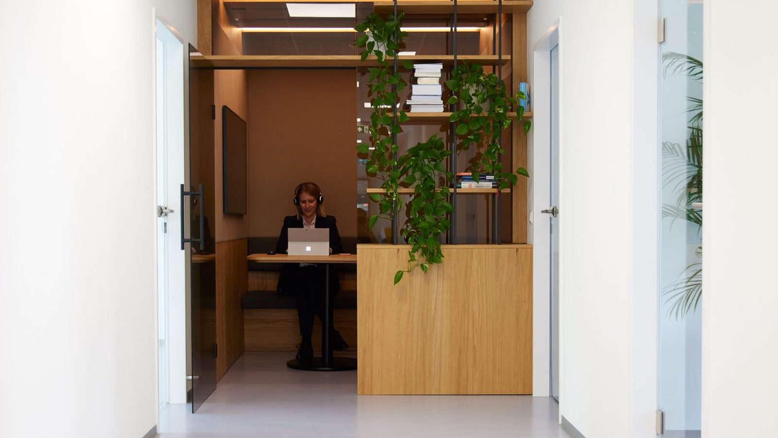 Blick durch einen hellen Bürogang bei ReqPOOL in eine Besprechungsnische aus Holz mit Pflanzen, in der eine Mitarbeiterin am Laptop arbeitet.