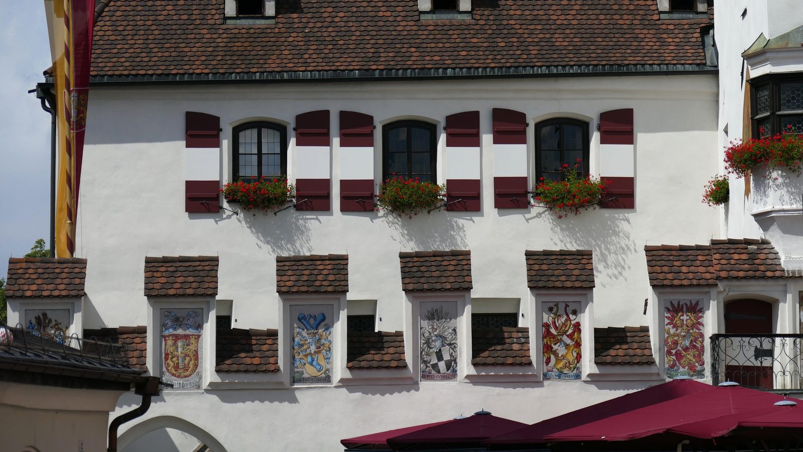 Fassade des Rathauses am Oberen Stadtplatz in Hall in Tirol mit dunkelroten Fensterläden, Blumenkästen und historischen Wappenfresken.