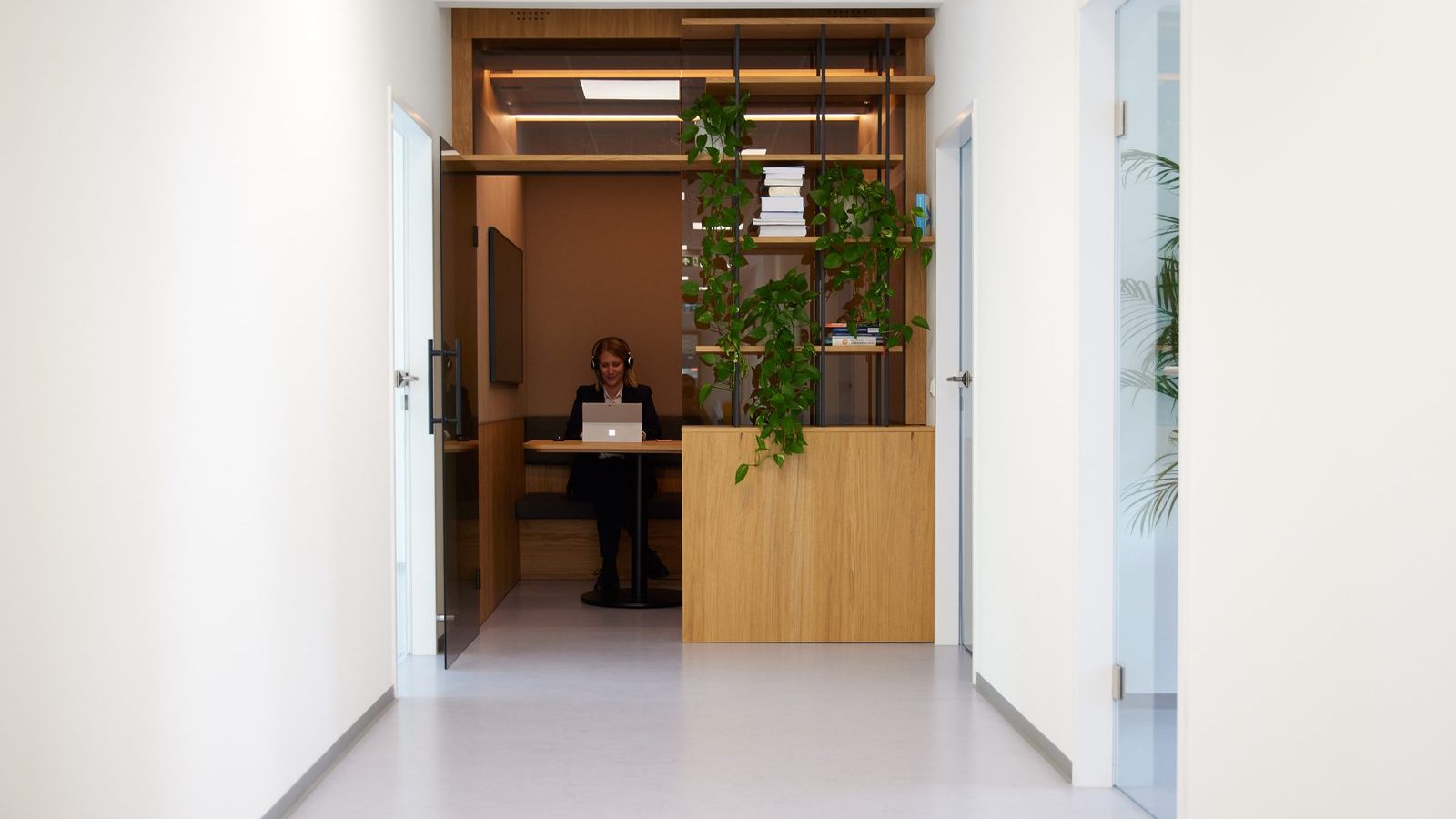 Blick durch einen hellen Bürogang von ReqPOOL auf eine Beraterin, die mit Kopfhörern in einer holzvertäfelten Nische am Laptop arbeitet.