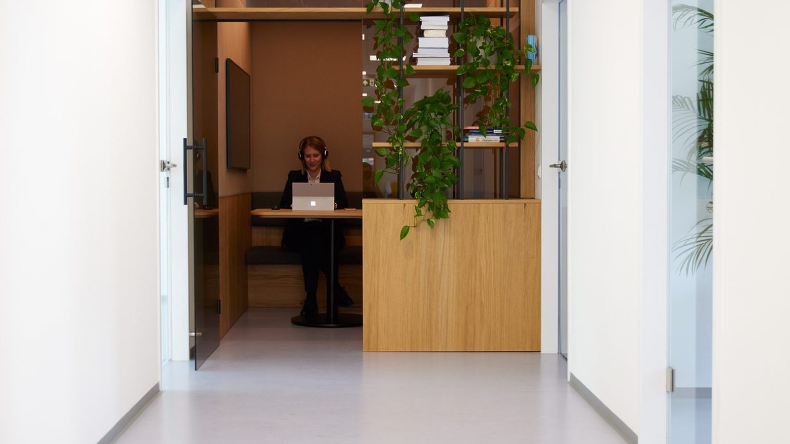 Heller Bürogang bei ReqPOOL, an dessen Ende eine Beraterin mit Kopfhörern in einer Holznische am Laptop arbeitet, daneben ein Regal mit Pflanzen und Büchern.