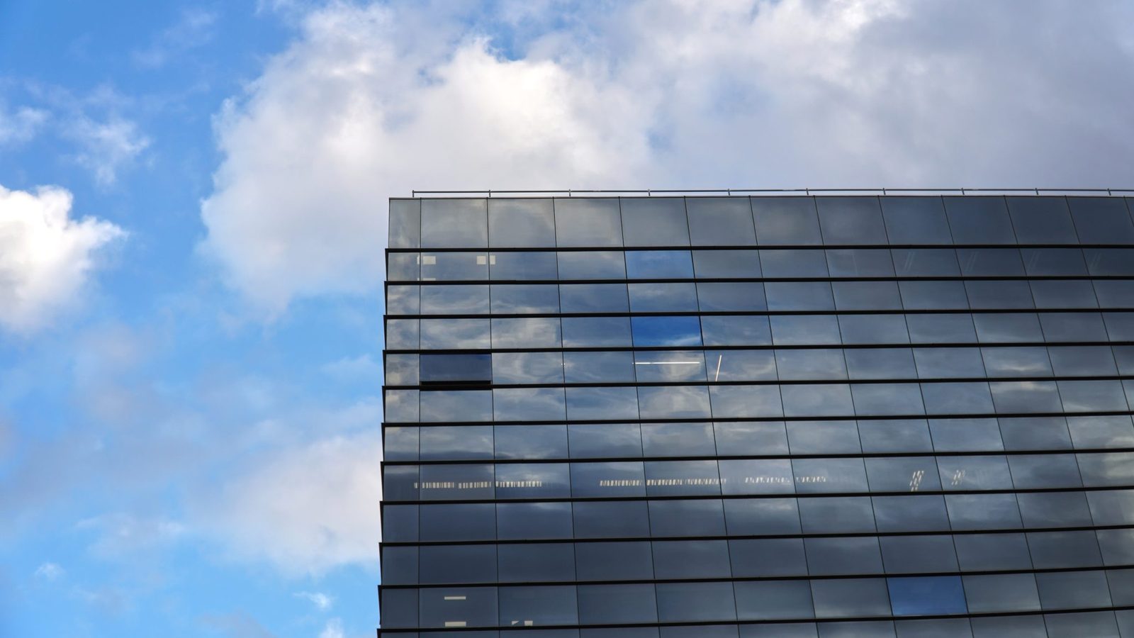 Glasfassade eines modernen Bürogebäudes von unten fotografiert, in deren Fenstern sich ein blauer Himmel mit weißen Wolken spiegelt.