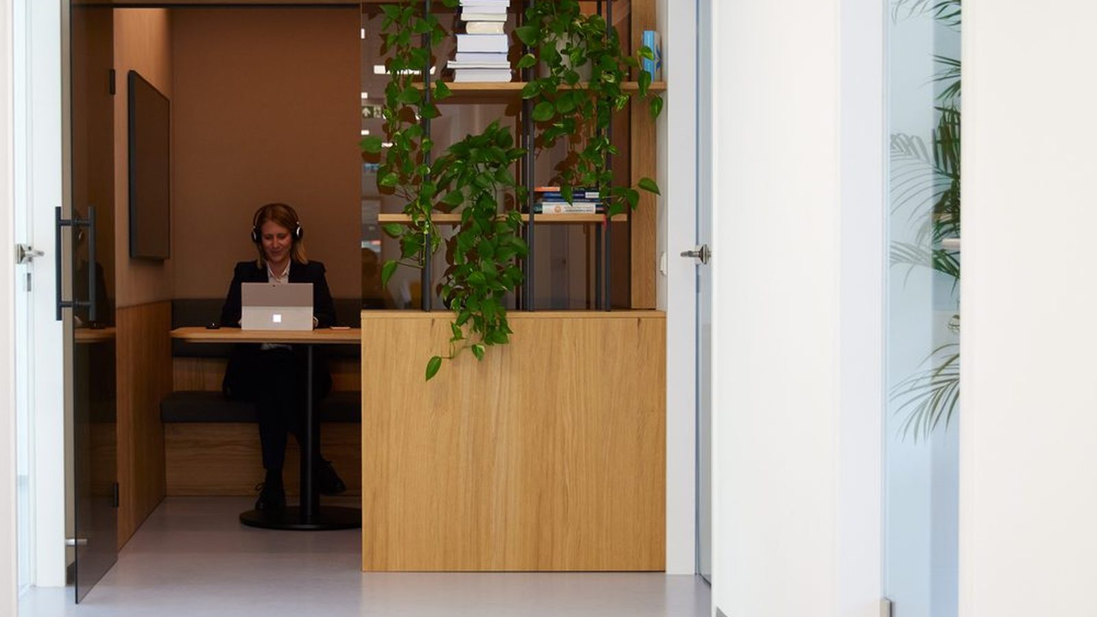 Heller Bürogang bei ReqPOOL, an dessen Ende eine Beraterin mit Kopfhörern in einer Holznische am Laptop arbeitet, daneben ein Regal mit Pflanzen und Büchern.