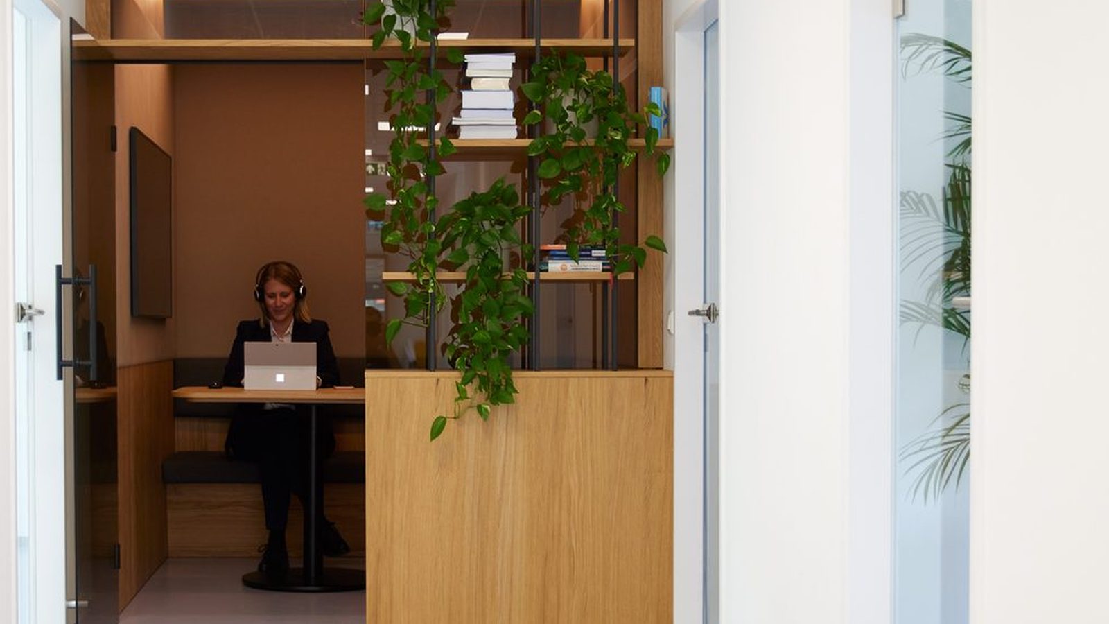 Heller Flur in einem ReqPOOL-Büro: Durch eine geöffnete Glastür arbeitet eine Beraterin mit Headset am Laptop in einer Holznische, daneben ein Regal mit Pflanzen.