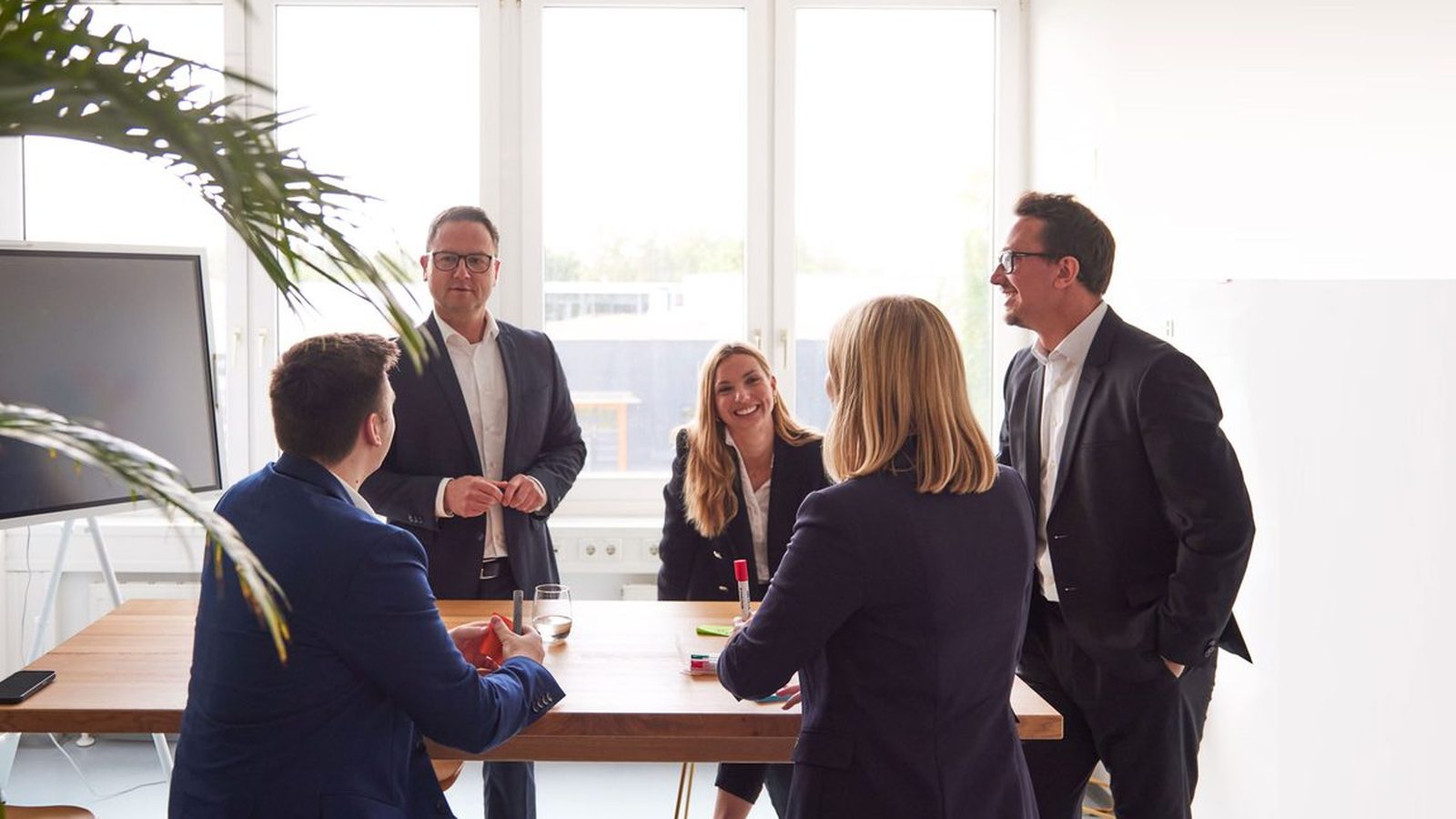 Fünf Beraterinnen und Berater von ReqPOOL besprechen sich an einem hohen Holztisch in einem hellen Büro mit Bildschirm und Zimmerpflanze.