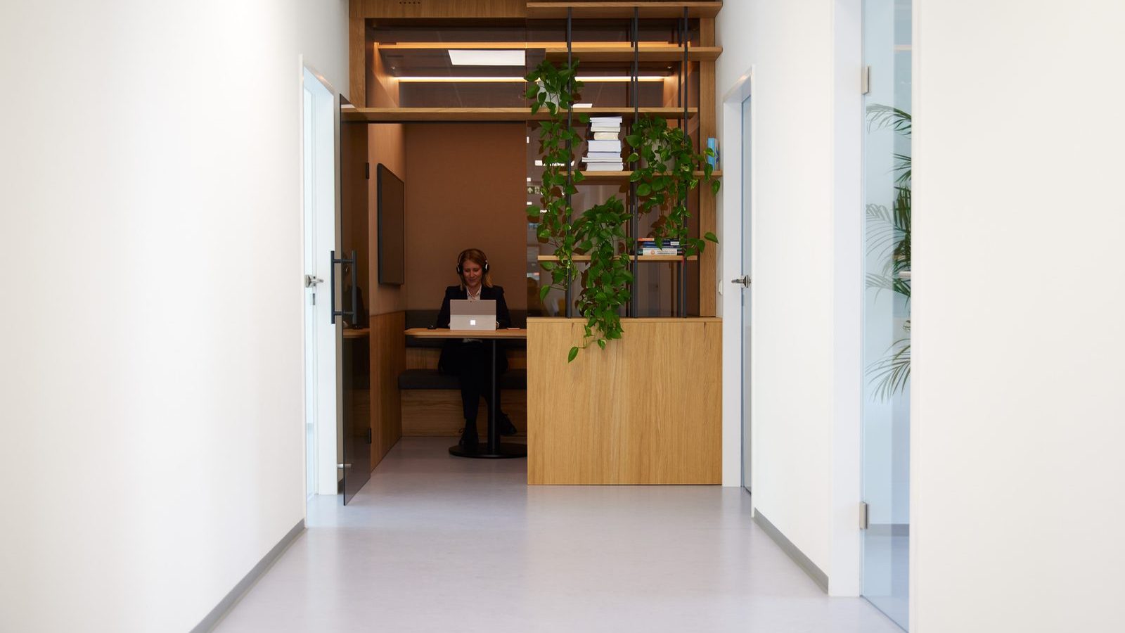 Heller Bürogang bei ReqPOOL, an dessen Ende eine Beraterin mit Kopfhörern in einer holzverkleideten Nische am Laptop arbeitet, daneben ein begrüntes Regal.