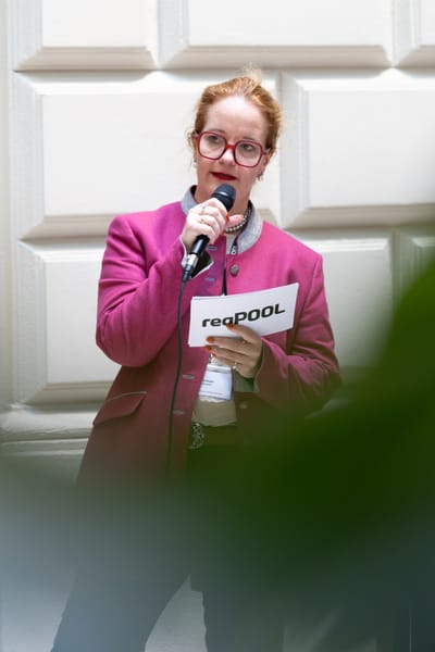 Rednerin in pinkem Blazer spricht mit Mikrofon vor heller Fassade