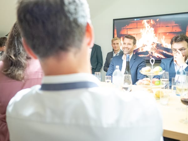 Gäste am Tisch mit Getränken und Obst, im Hintergrund ein Kaminfeuer auf dem Bildschirm