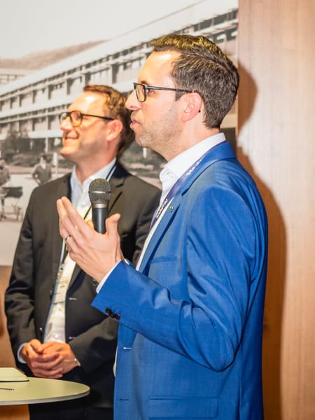Redner im blauen Sakko mit Mikrofon vor einer Wand mit Schwarzweißfoto