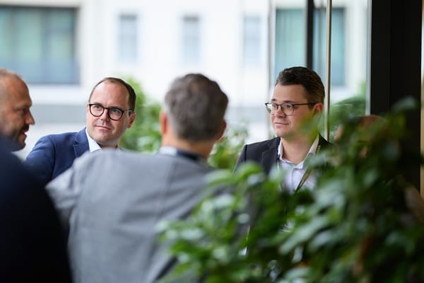 Zwei Gäste hören einem Gespräch vor großen Fenstern und einer Zimmerpflanze zu