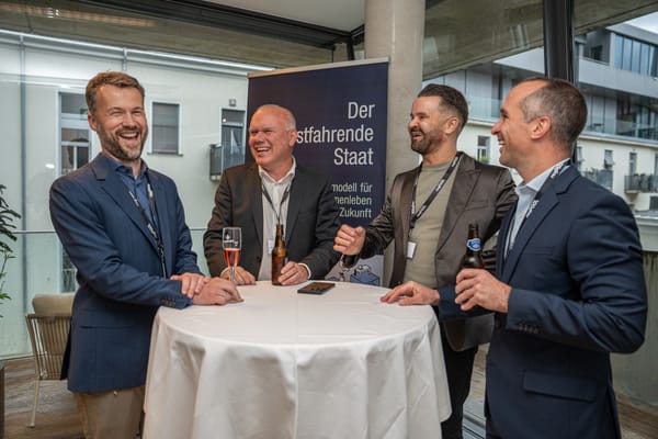Vier Männer lachen an einem Stehtisch auf der Terrasse, dahinter ein Banner zum „selbstfahrenden Staat“